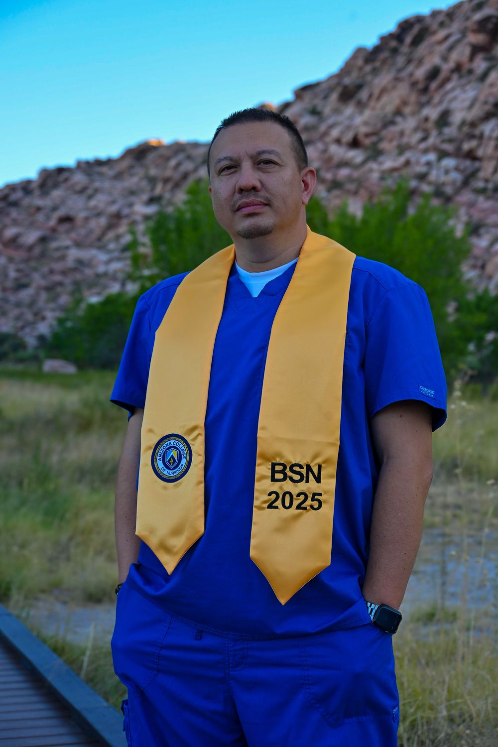 Nursing graduate in blue scrubs wearing gold BSN 2025 stole poses seriously against rocky landscape.