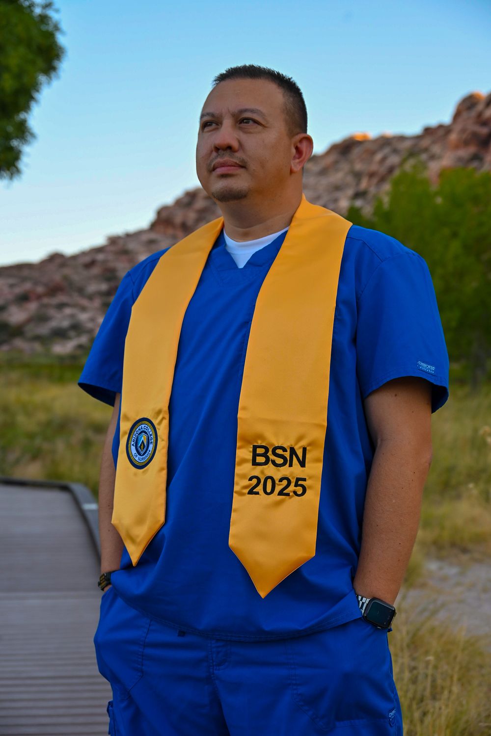 Nursing graduate wearing gold BSN 2025 stole in blue scrubs stands on outdoor boardwalk.