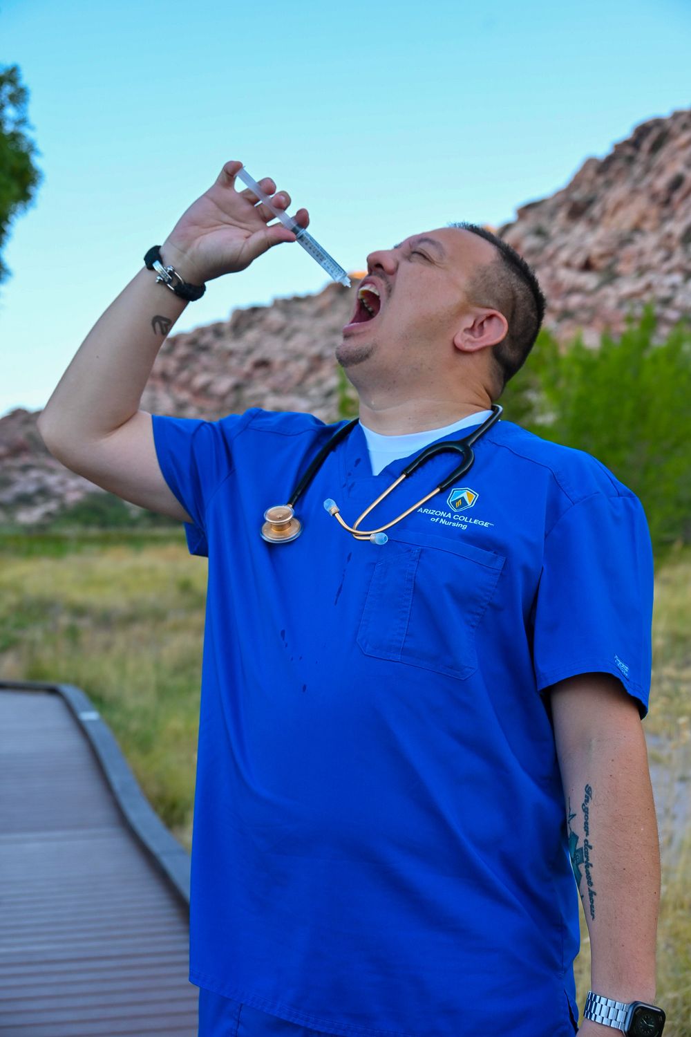 Male nurse in blue scrubs humorously pretends to drink from a syringe outdoors.