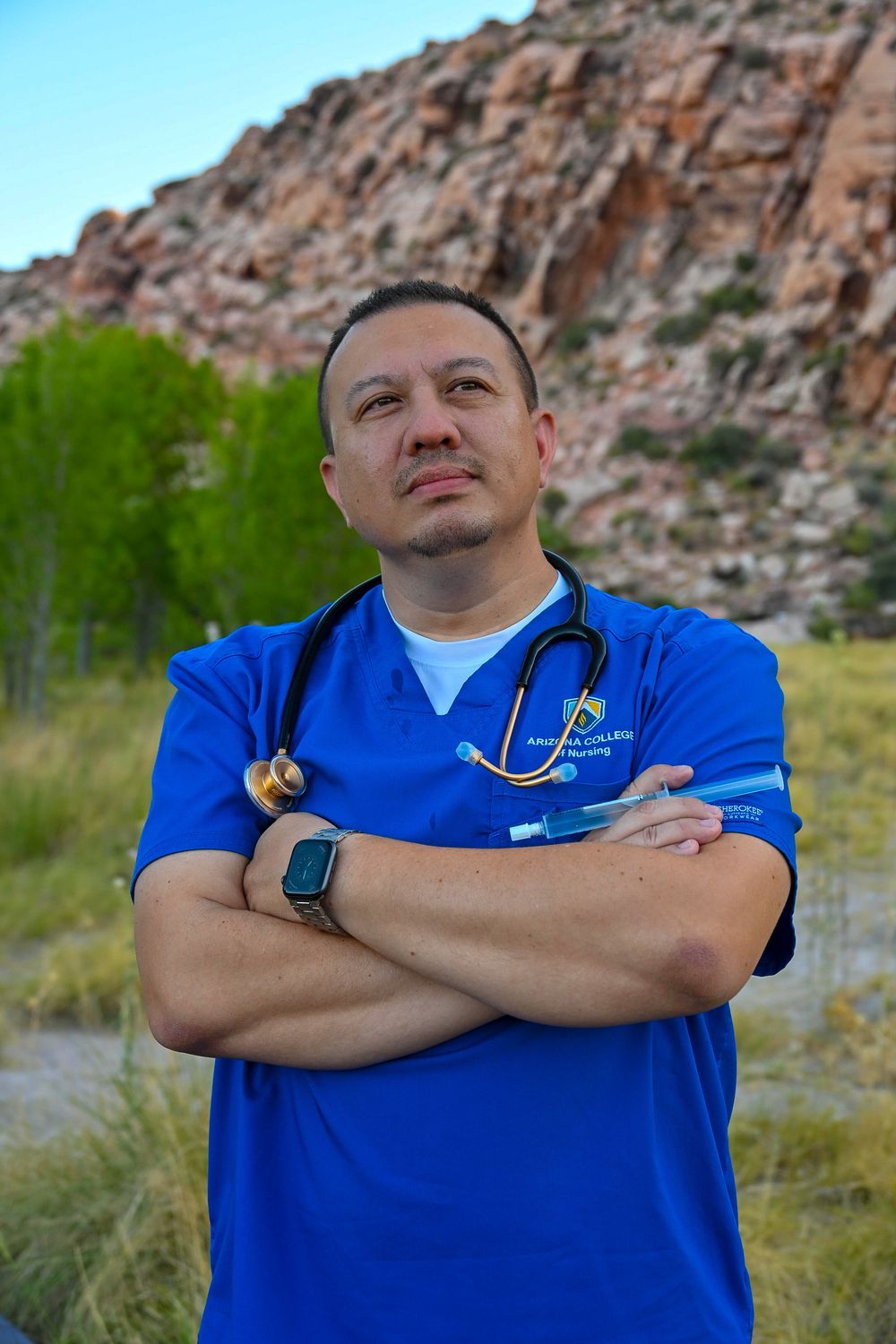 Male nurse in blue scrubs with stethoscope stands arms crossed near red rock landscape.