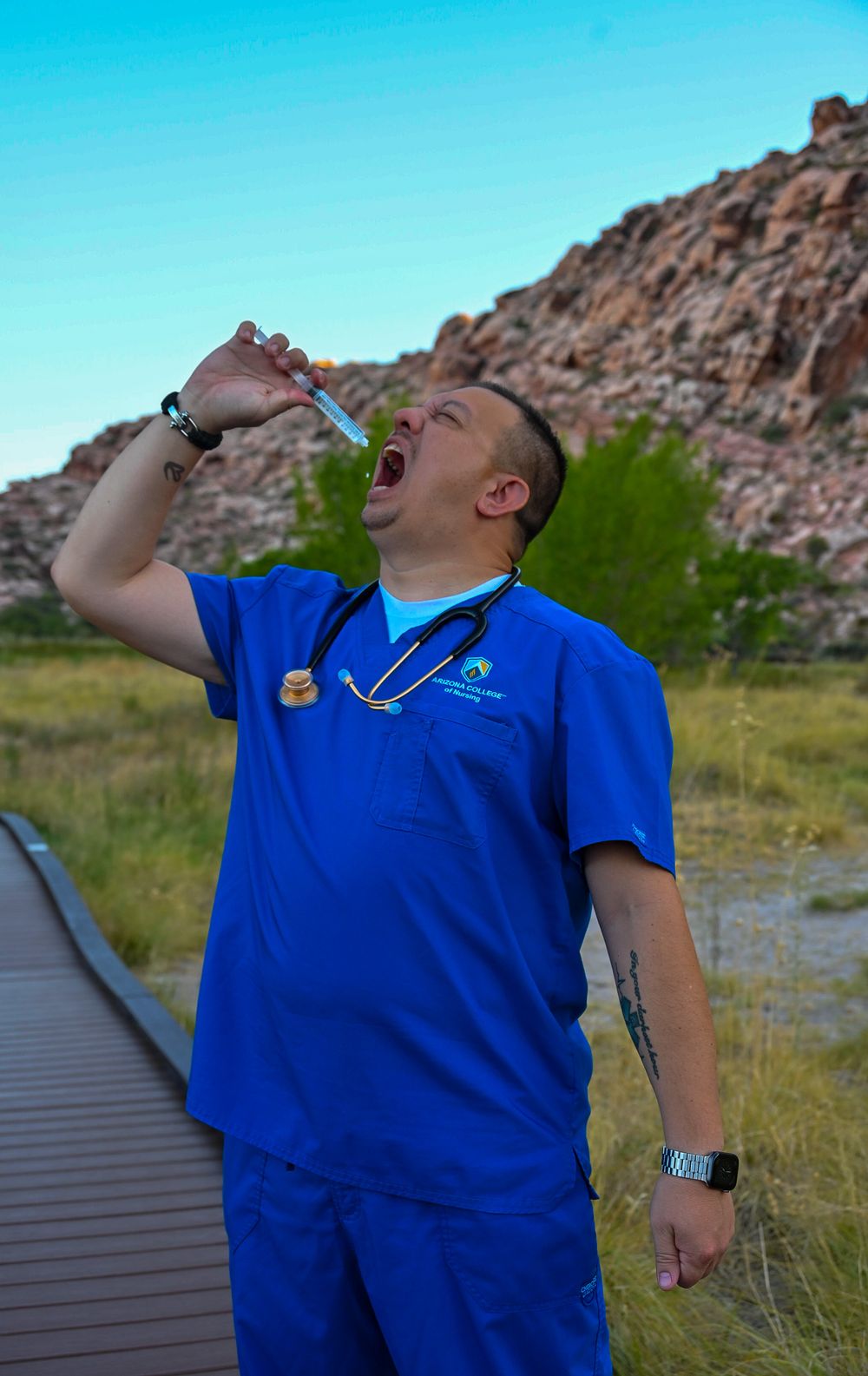 Nursing graduate in blue scrubs humorously pretends to drink from syringe outdoors near red rocks.