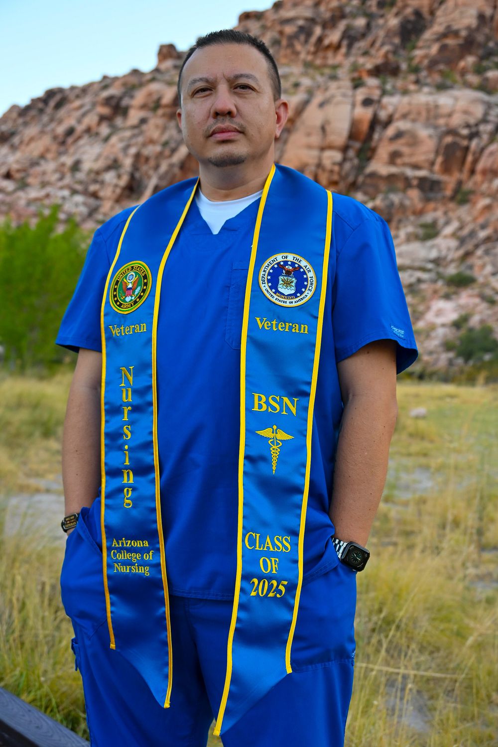Veteran BSN nursing graduate in blue scrubs with blue and gold stole stands in desert landscape for portrait.