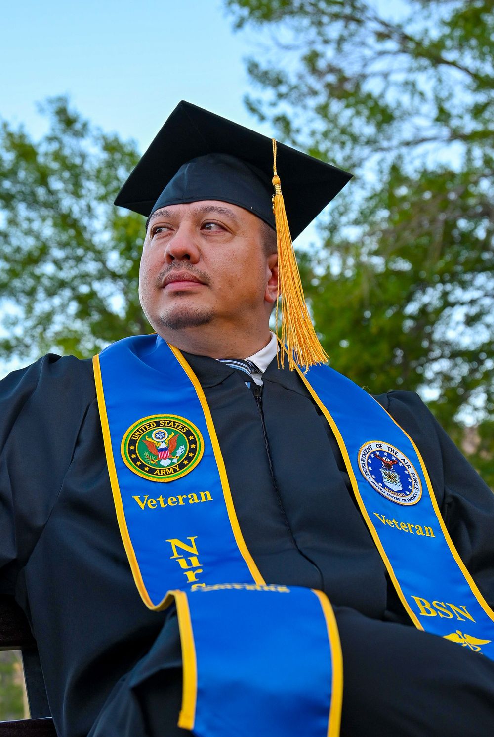 Veteran BSN nursing graduate in black cap and gown with blue stole sits outdoors looking upward confidently.