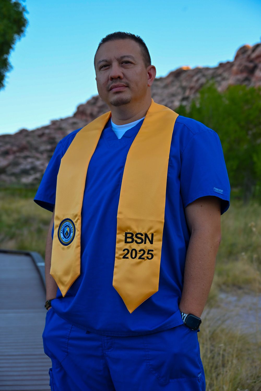 Man in blue nursing scrubs with gold BSN 2025 graduation stole stands on boardwalk in desert landscape.