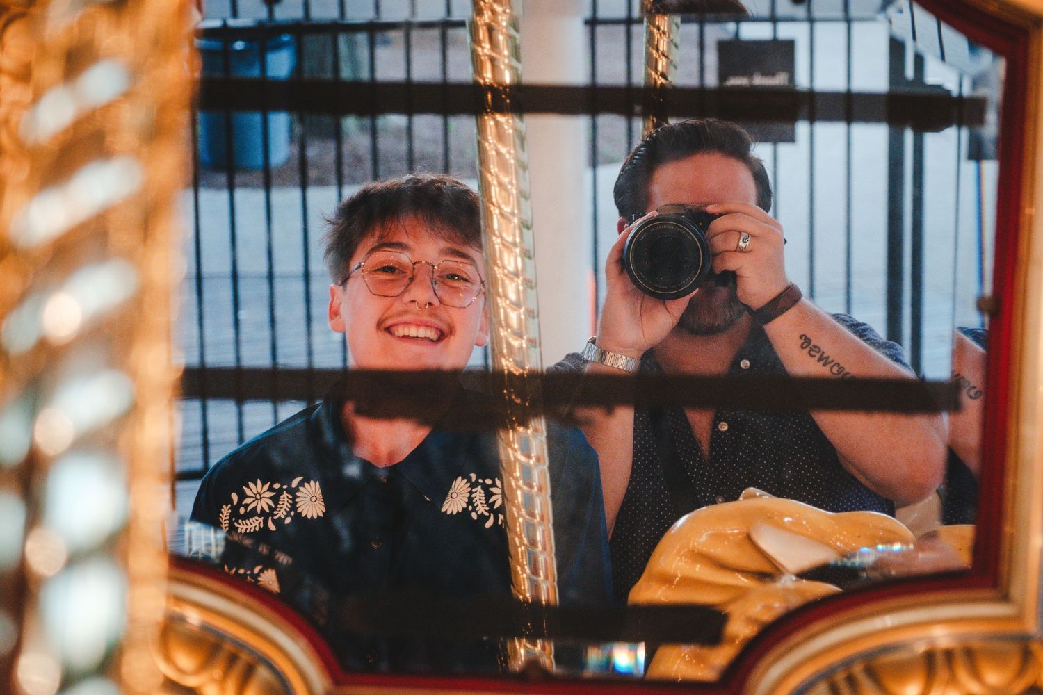 Two friends sit on a carousel at dusk, one smiling while the other holds a camera beside orange-tinted rope lights.
