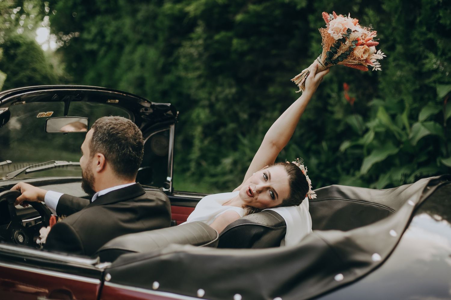 A couple celebrates in a vintage convertible car as someone holds up a wedding bouquet against a green forest backdrop.