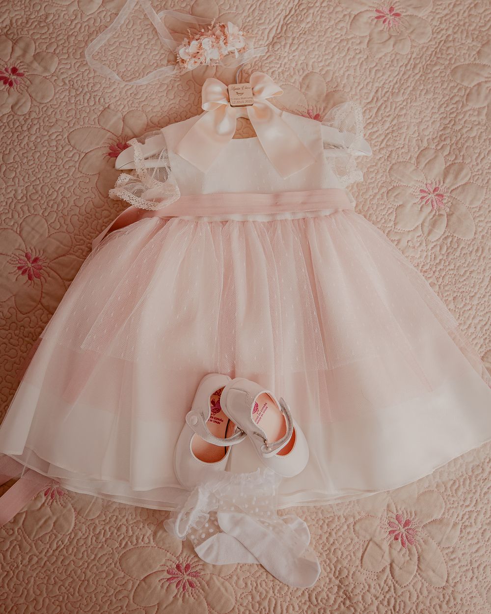 Delicate pink baby dress with ruffled layers and floral embroidery displayed on textured fabric background.