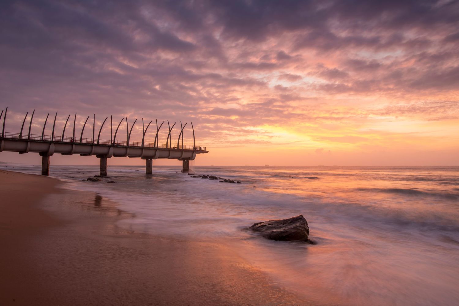 A wooden pier extends over the ocean at sunrise with dramatic pink clouds and gentle waves washing onto sandy beach.
