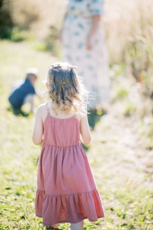 Small child in flowing pink tiered dress standing in sunny meadow with sunlit hair blowing in breeze.