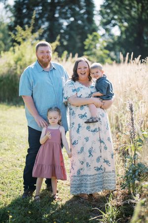 A family stands together in a sunny field with tall grass and wildflowers during a summer photo session.