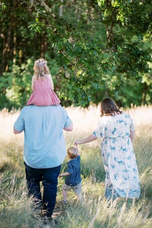 A family casually walks together through a grass field under tree branches on a sunny day.