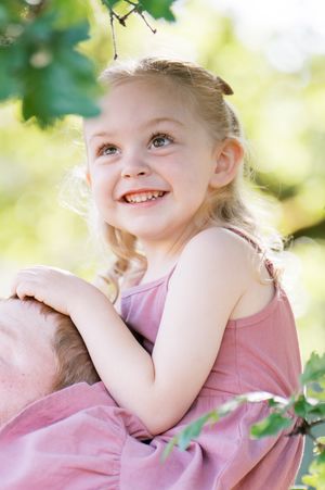 A child wearing pink smiles joyfully while playing outdoors on a bright sunny day with blurred greenery in the background.