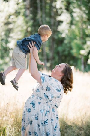 A tender moment captured outdoors as a woman in a floral dress lifts a small child in the air against a sunlit natural backdrop.