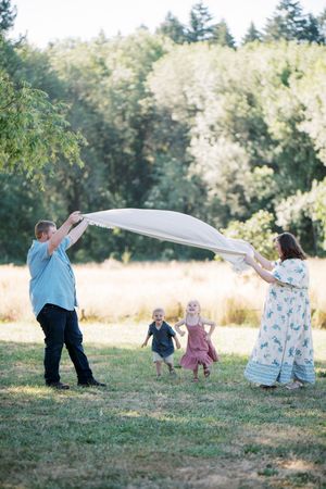 A group plays joyfully in a sunny field with a blanket while two children run between them in the grass.