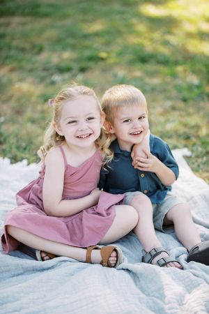 Two young children sit on a blanket outdoors while laughing and playing together in matching pink outfits.