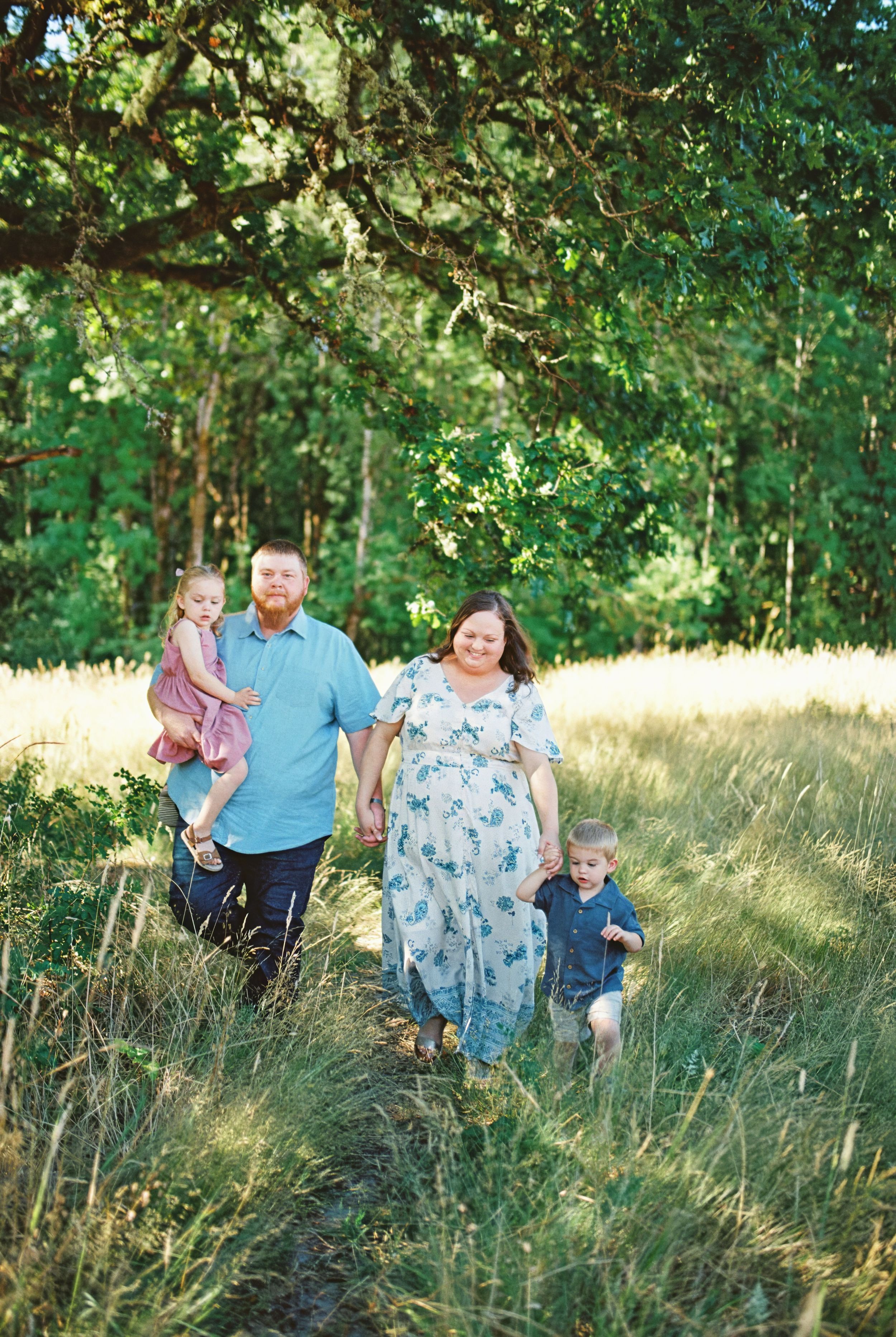 A family stands together in a sunlit meadow surrounded by trees and tall grass on a summer afternoon.