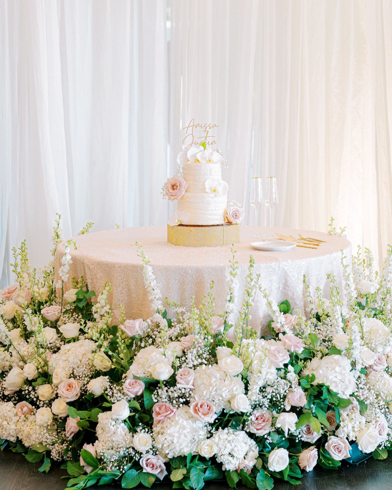 Elegant white wedding cake displayed on round table surrounded by lush white and pink floral arrangements.