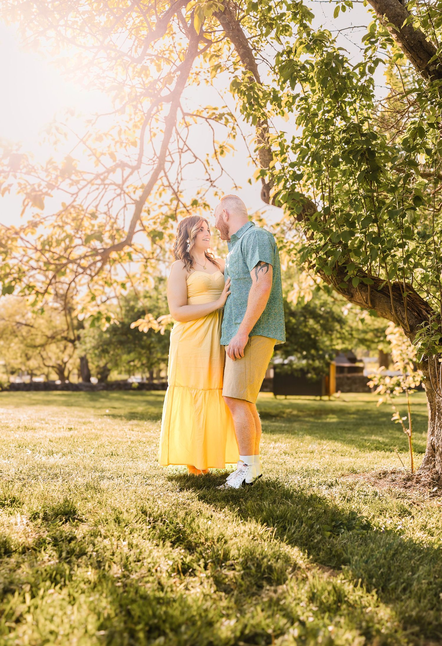 A couple shares a romantic moment under a sunlit tree in a park, with golden afternoon light filtering through the leaves.