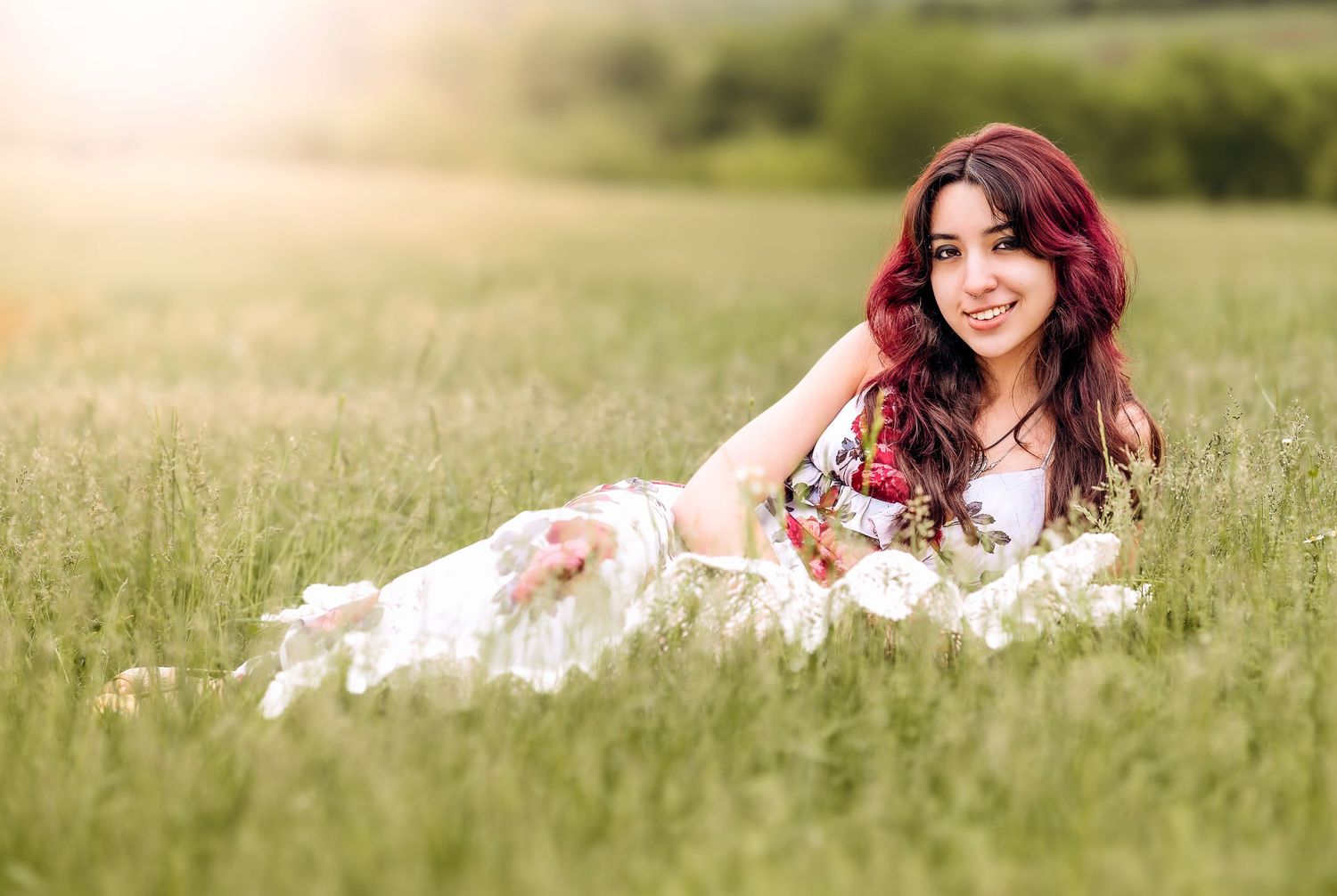 Woman with vibrant red hair lying in a sunny meadow wearing a white lace dress among tall grass.