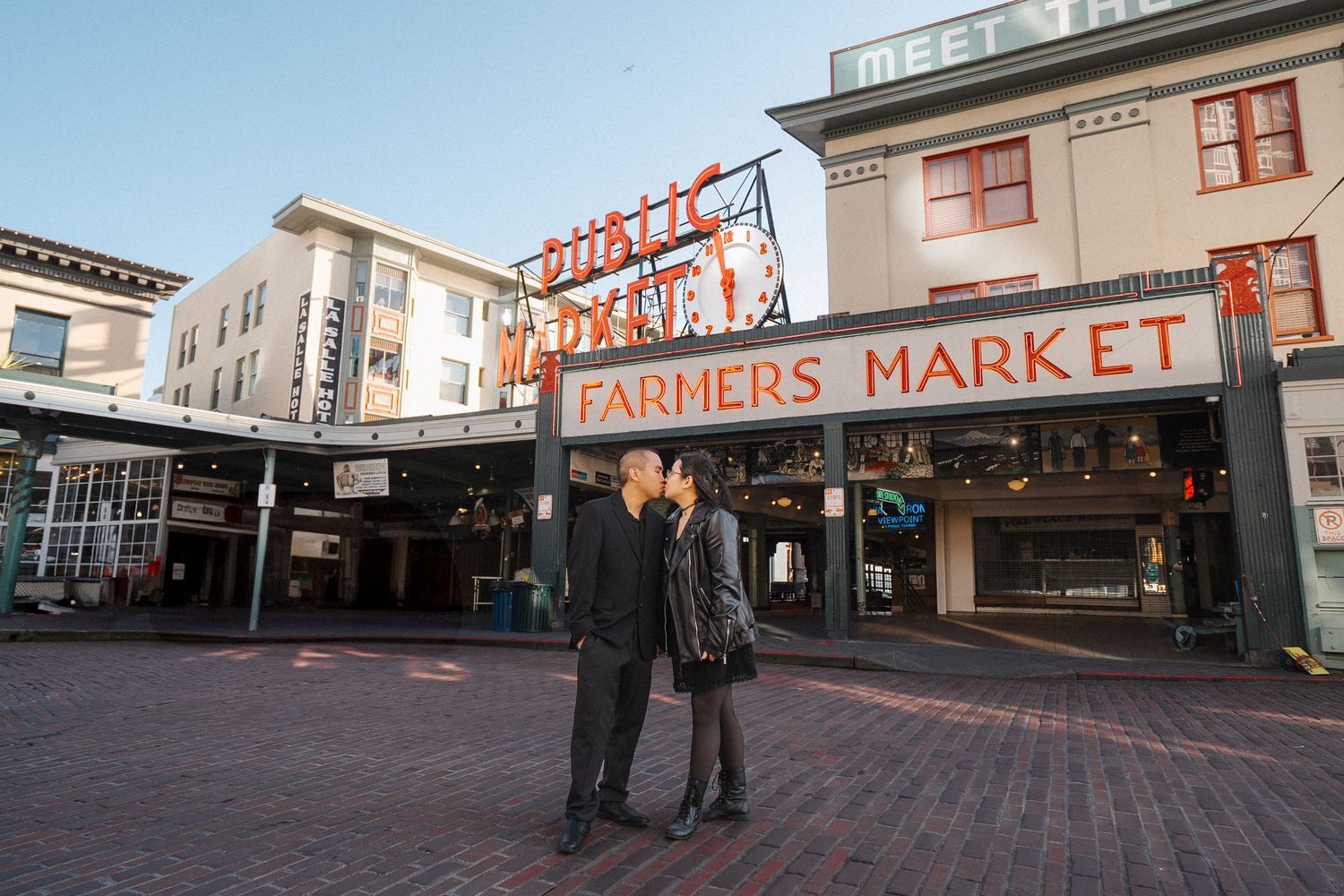 Two people stand on the brick pavement in front of the iconic Pike Place Market public sign in downtown Seattle.
