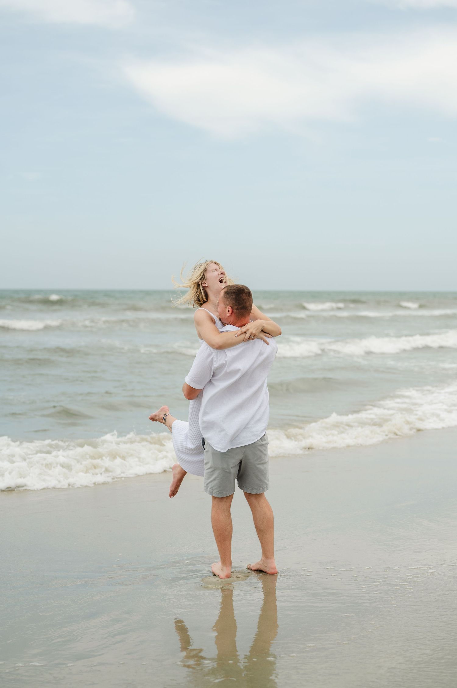 Romantic couple embracing on North Carolina beach with waves and cloudy sky in background.