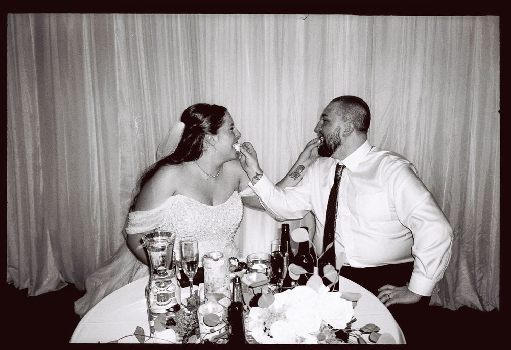 Black and white film photo of couple feeding each other local 88 donuts instead of wedding cake at their reception in southern Maine.