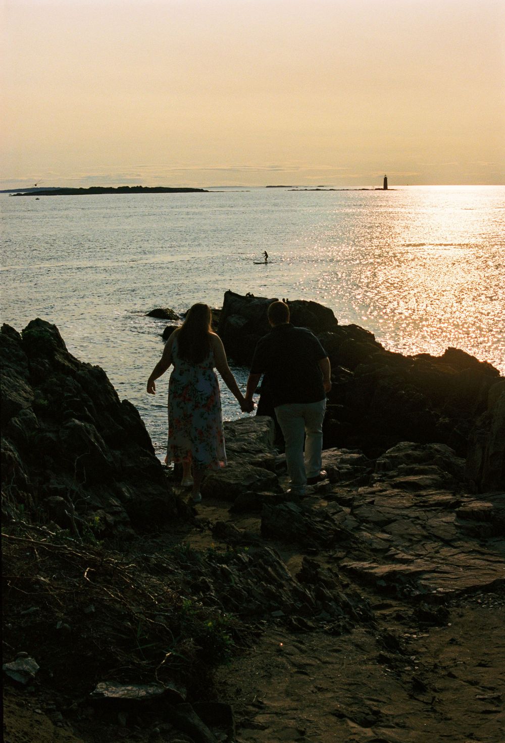 Couple holds hands on the rocks by the beach in the early morning sunshine with the light reflecting off the water on their engagement shoot