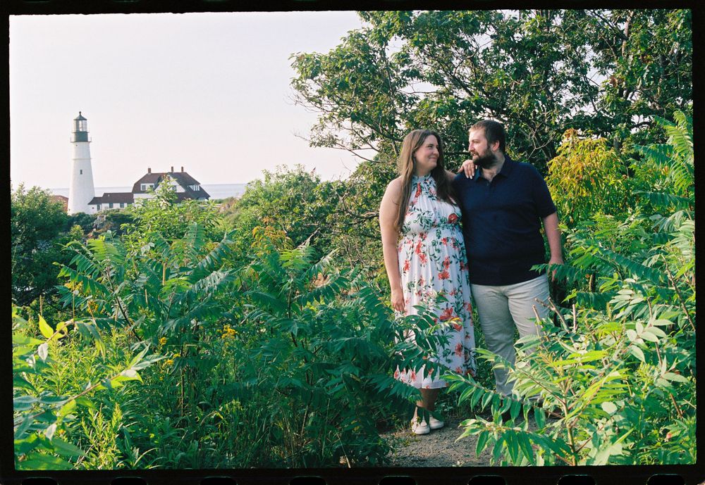 Engaged couple smiles at each other surrounded by lush greenery with a lighthouse in the background at Fort Williams Park in Cape Elizabeth.