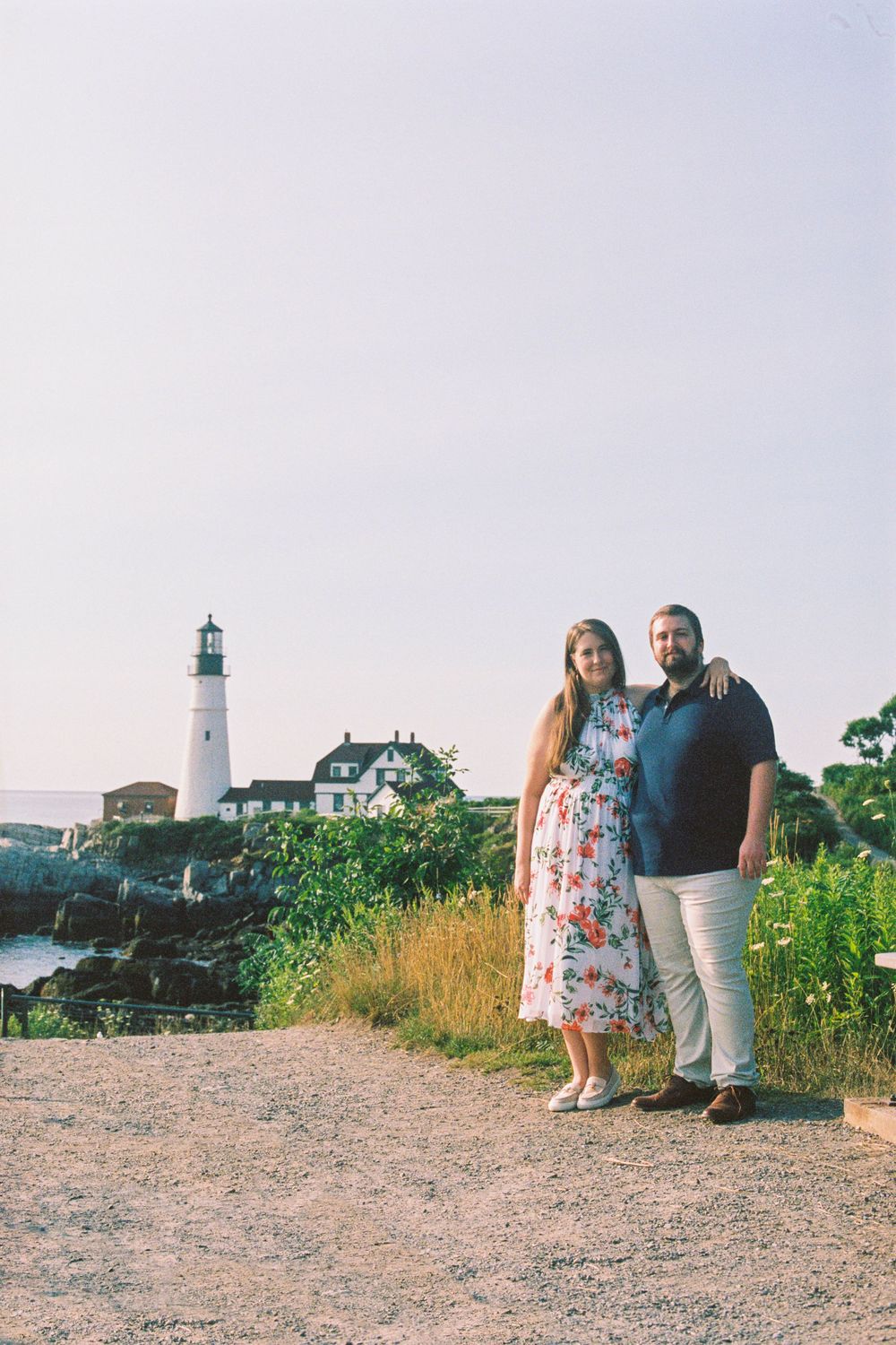 Couple poses sweetly together in summer clothing by the lighthouse in Fort Williams park in Cape Elizabeth, Maine on color film.