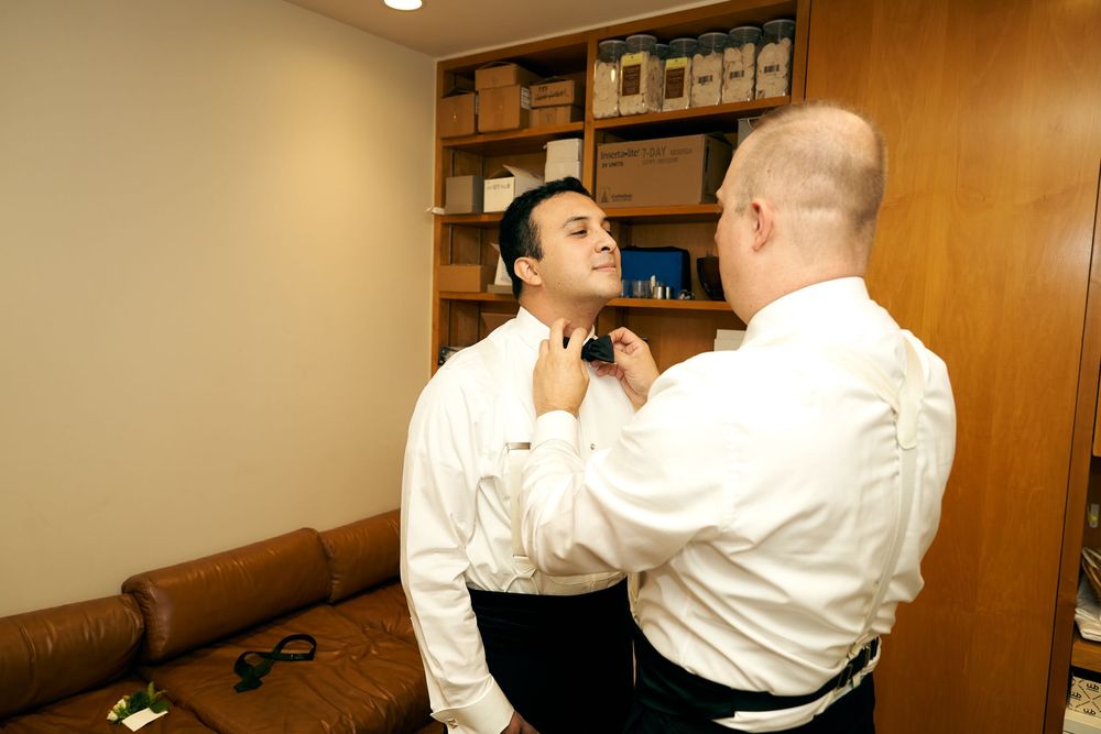 Two people in formal attire adjusting clothing in a room with wooden cabinets. Siant peters church