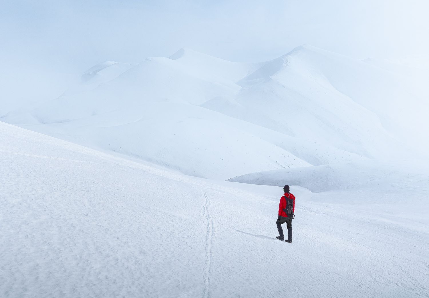 Hiker in red jacket walking across snow-covered mountain slopes under a pale winter sky