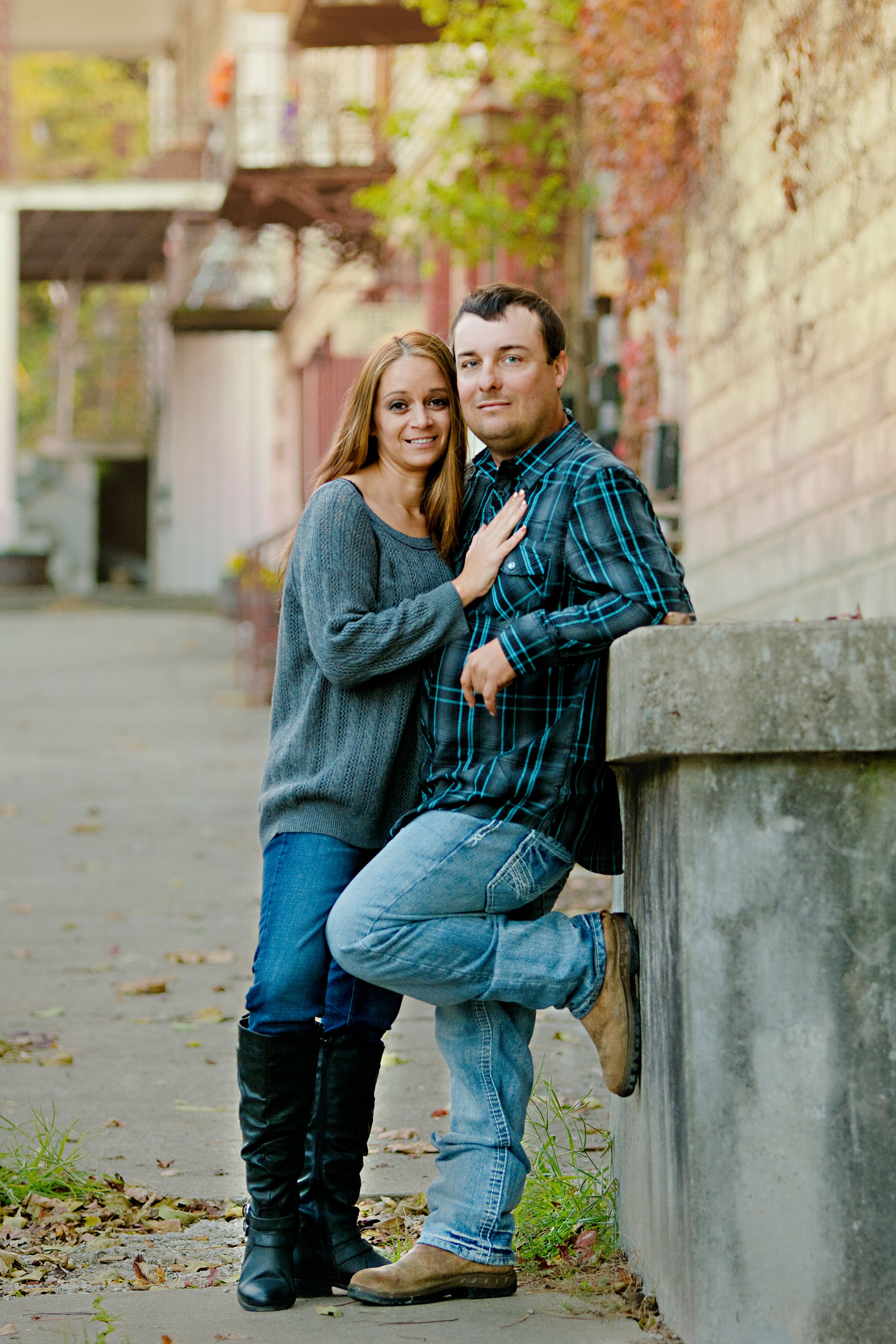 A couple in casual fall attire poses together against a brick wall on an urban sidewalk during an engagement shoot.