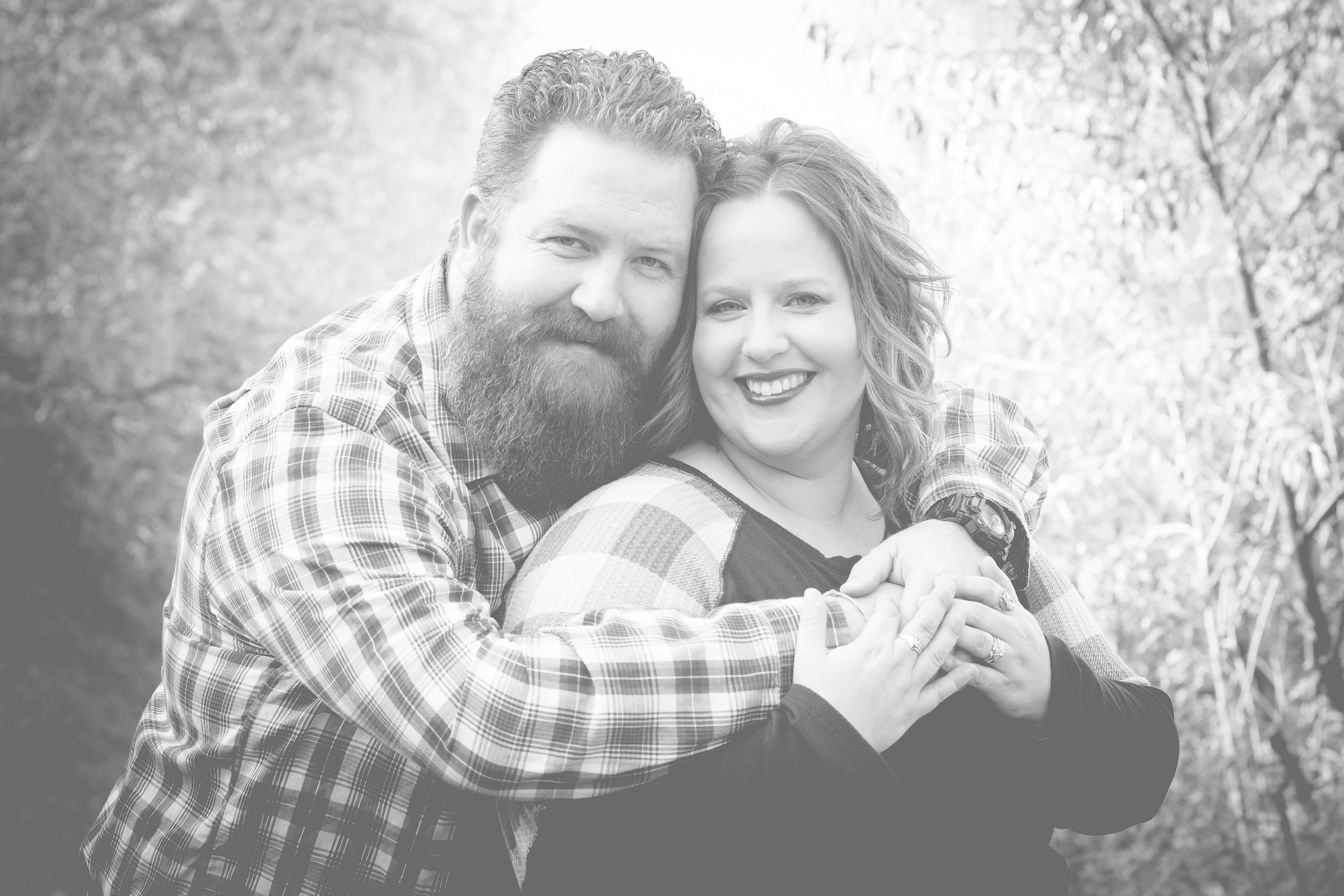 A loving couple in a black and white photo embraces outdoors, with one wearing a plaid shirt and sporting a full beard.