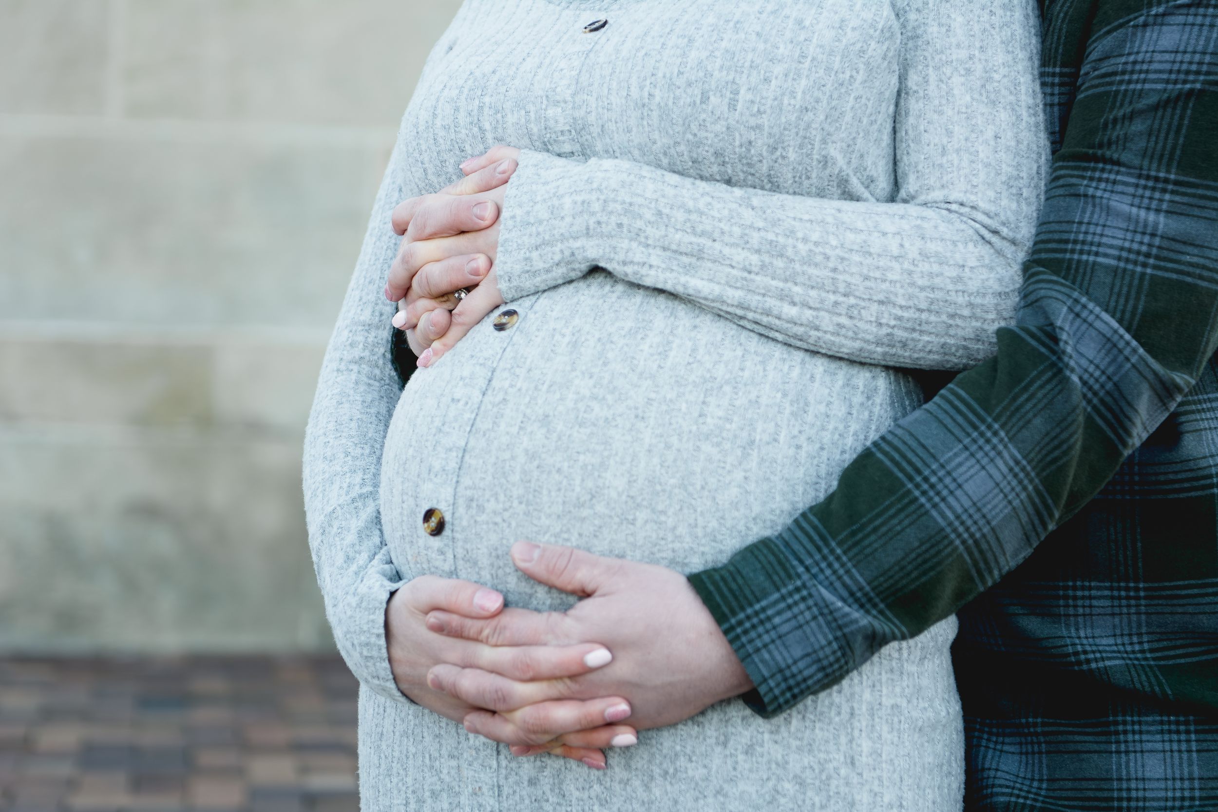A pregnant woman in a gray sweater being lovingly embraced from behind with hands resting on her belly.