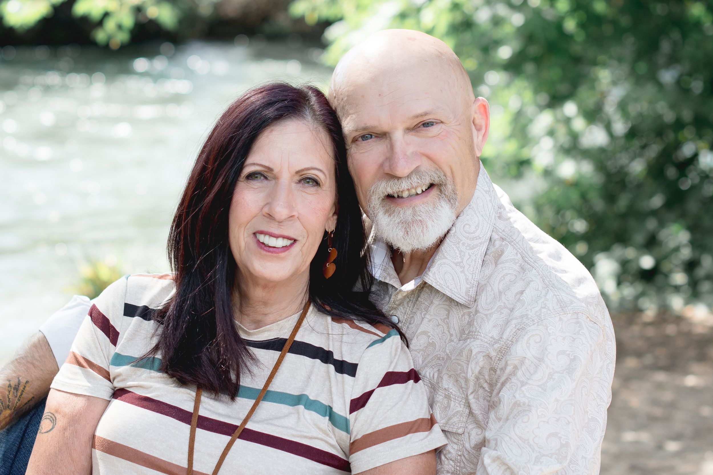 A couple smiles at the camera while sitting outdoors with greenery and water in the background.