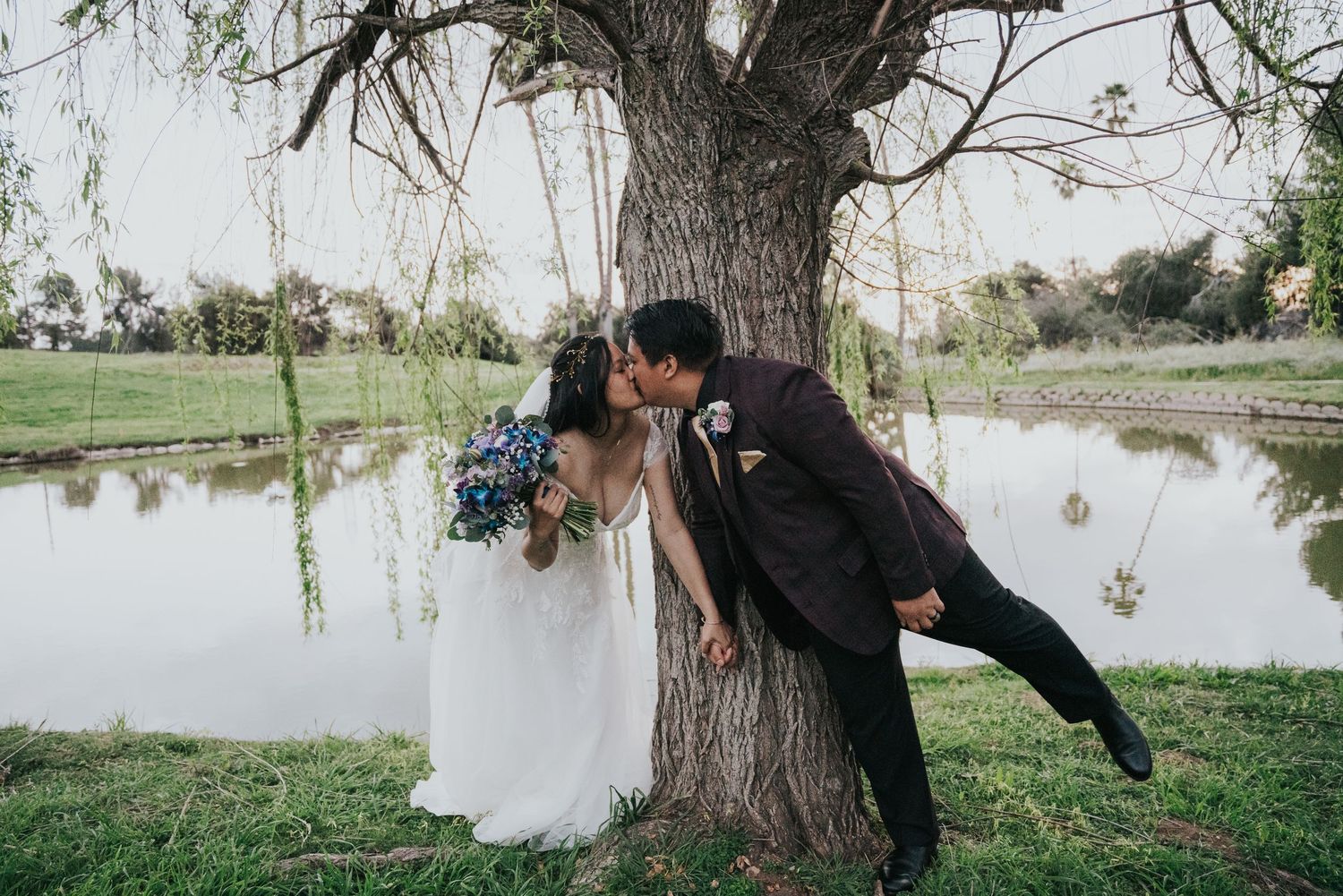 A couple in wedding attire share a romantic moment by a willow tree near a serene pond.