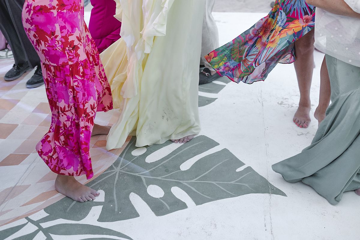Colorful floral dresses worn by barefoot women dancing on a white floor with a monstera leaf design.