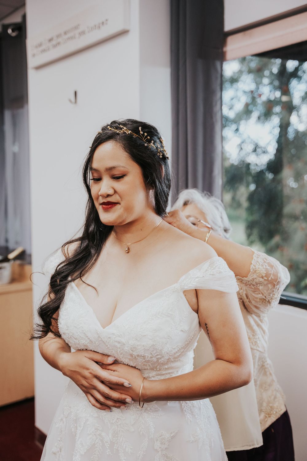 A bride in an off-shoulder lace wedding gown smiles while getting ready, wearing a delicate hair accessory.