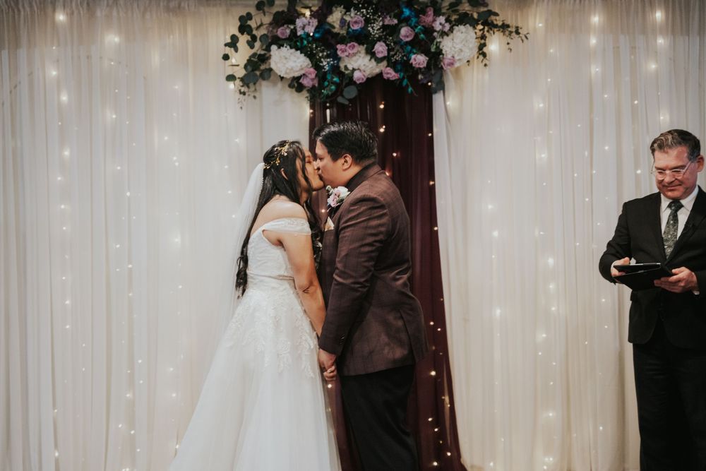 A couple shares their first kiss during a wedding ceremony under an arch with white drapery and floral decorations.