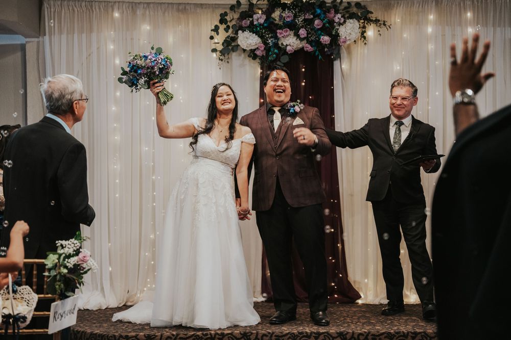 Newlyweds celebrate joyfully at wedding ceremony under floral arch with elegant white curtain backdrop.