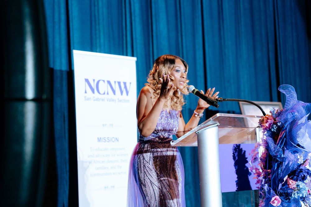 A speaker at a podium addresses an NCNW event while wearing a flowing light-colored dress on a blue-lit stage.