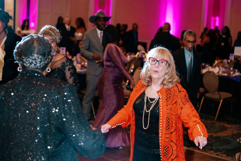 A lively dance floor scene illuminated by pink lights with a person in a bright orange outfit dancing among the crowd.