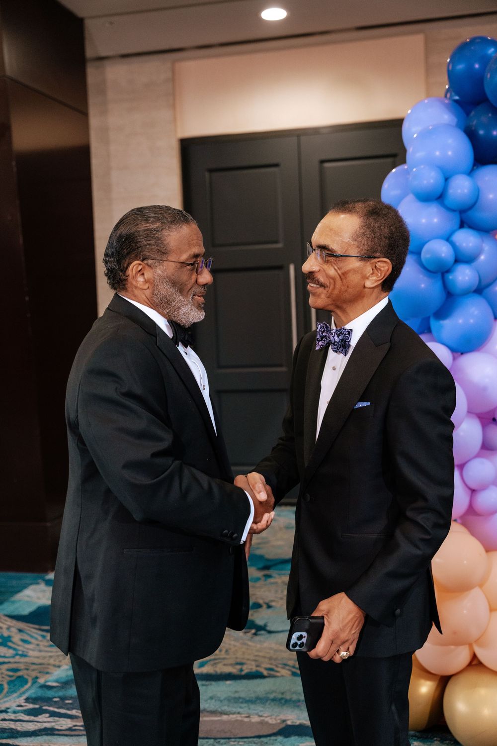 Two men dressed in formal suits converse and laugh against a backdrop of blue and peach colored balloons.