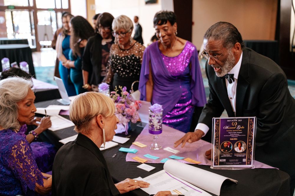 Attendees in purple formal attire gather around a table at a social event looking at documents together.