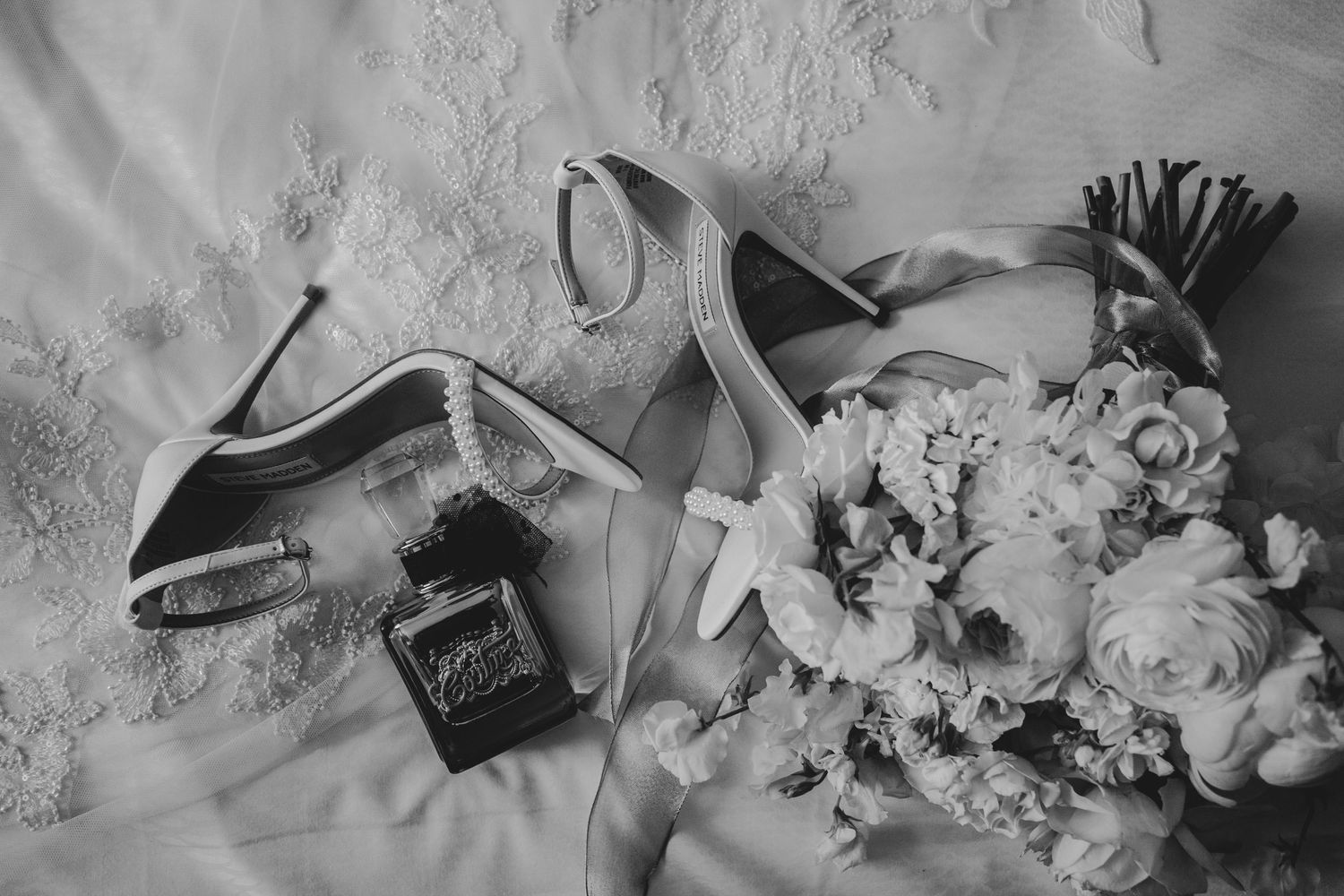 Black and white photo of elegant wedding accessories including shoes, rings, and a delicate floral bouquet.
