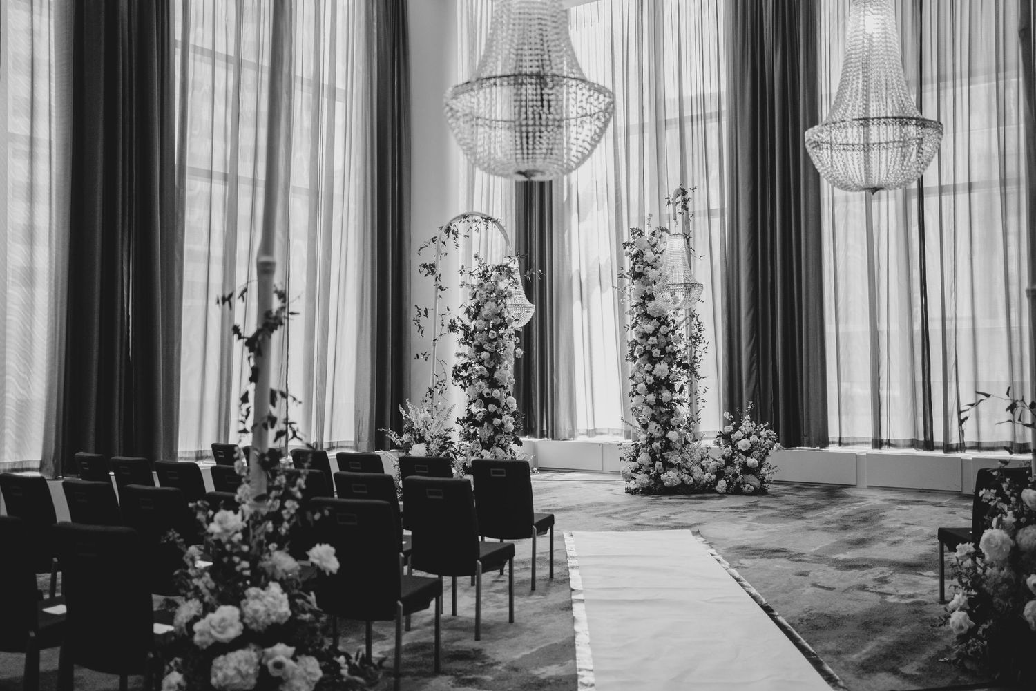 Black and white interior photo of an elegant wedding ceremony venue with floral arrangements and chandeliers.