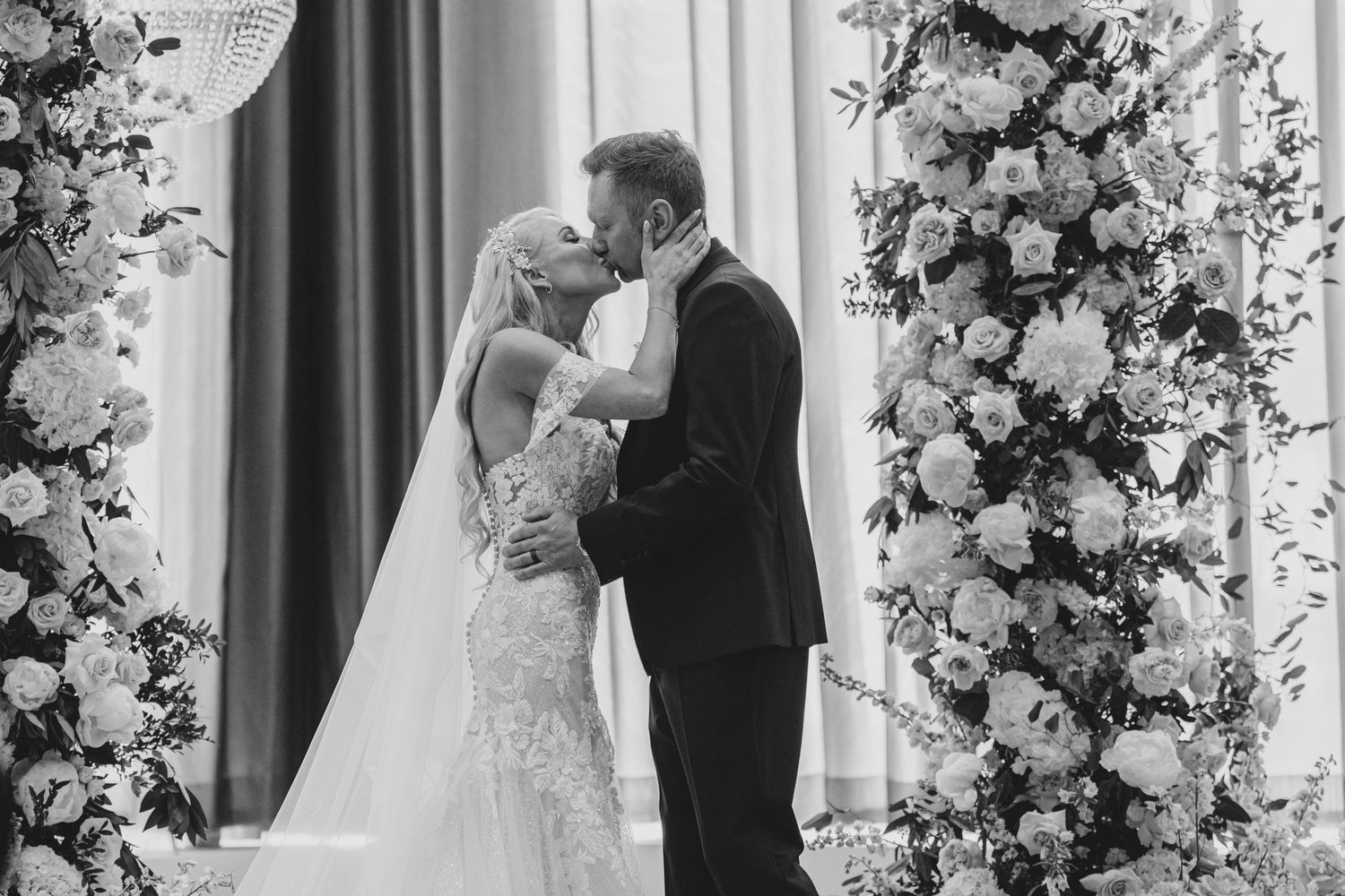 A wedding couple shares a kiss under elegant floral arches decorated with white flowers and draping.