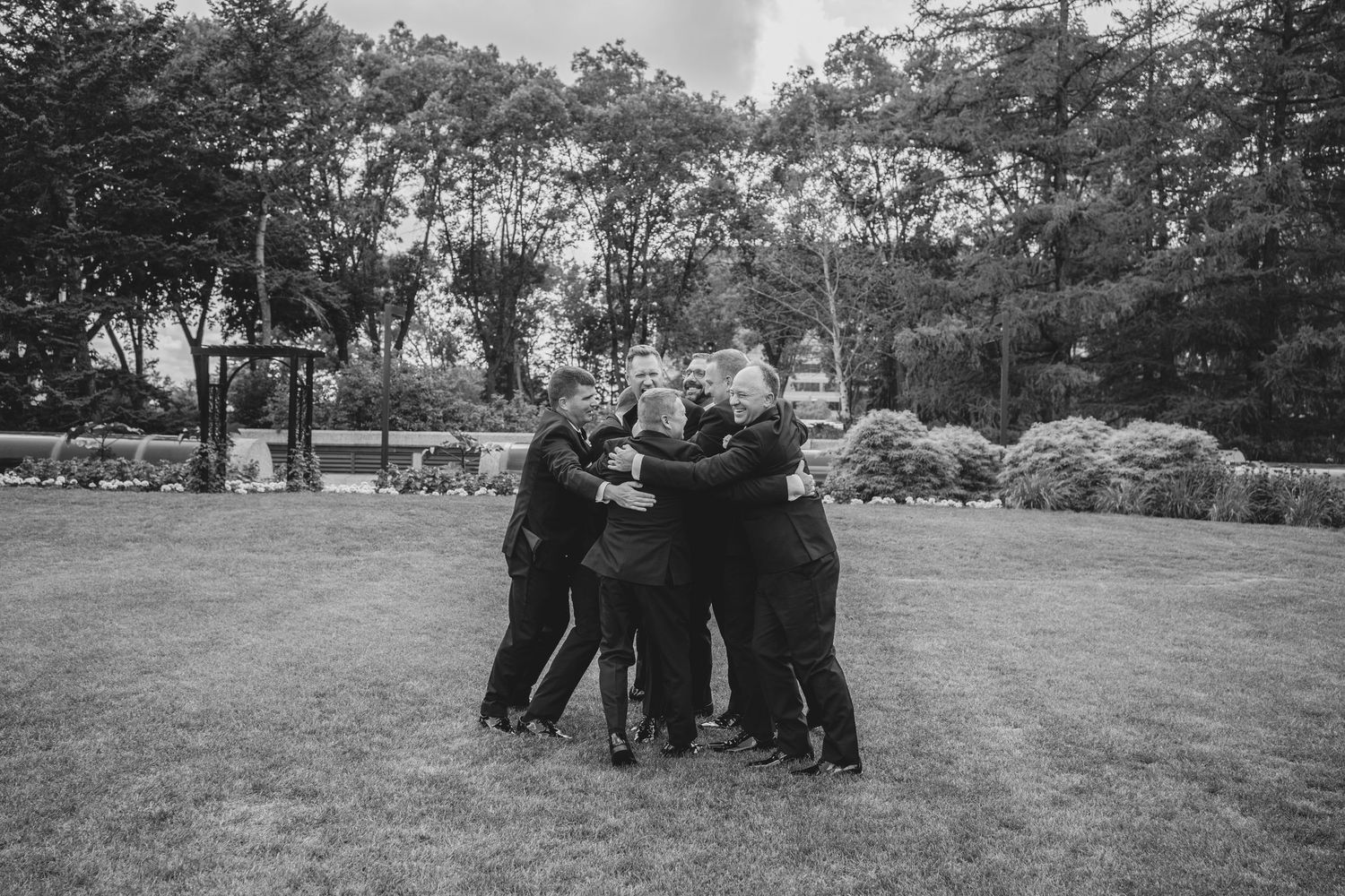 A group in formal attire stands embracing in a circle on a grassy lawn surrounded by trees in black and white.