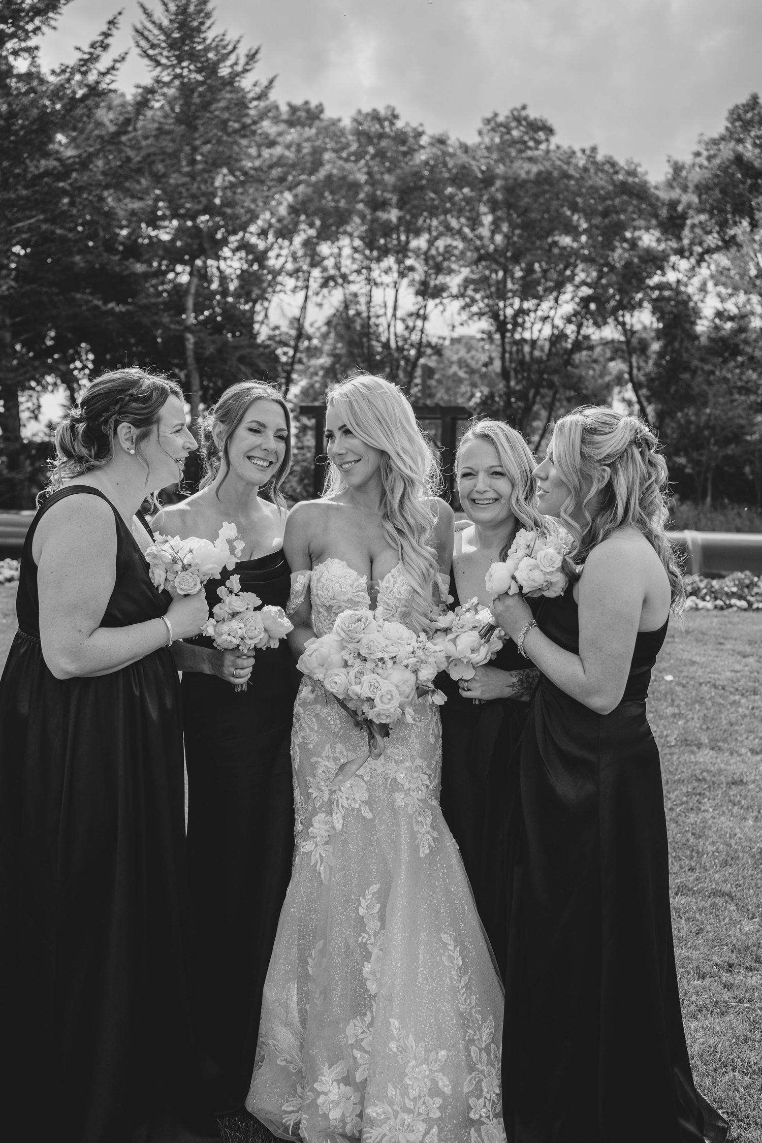 Wedding party in black dresses and a white gown pose together outdoors with bouquets against a natural backdrop.