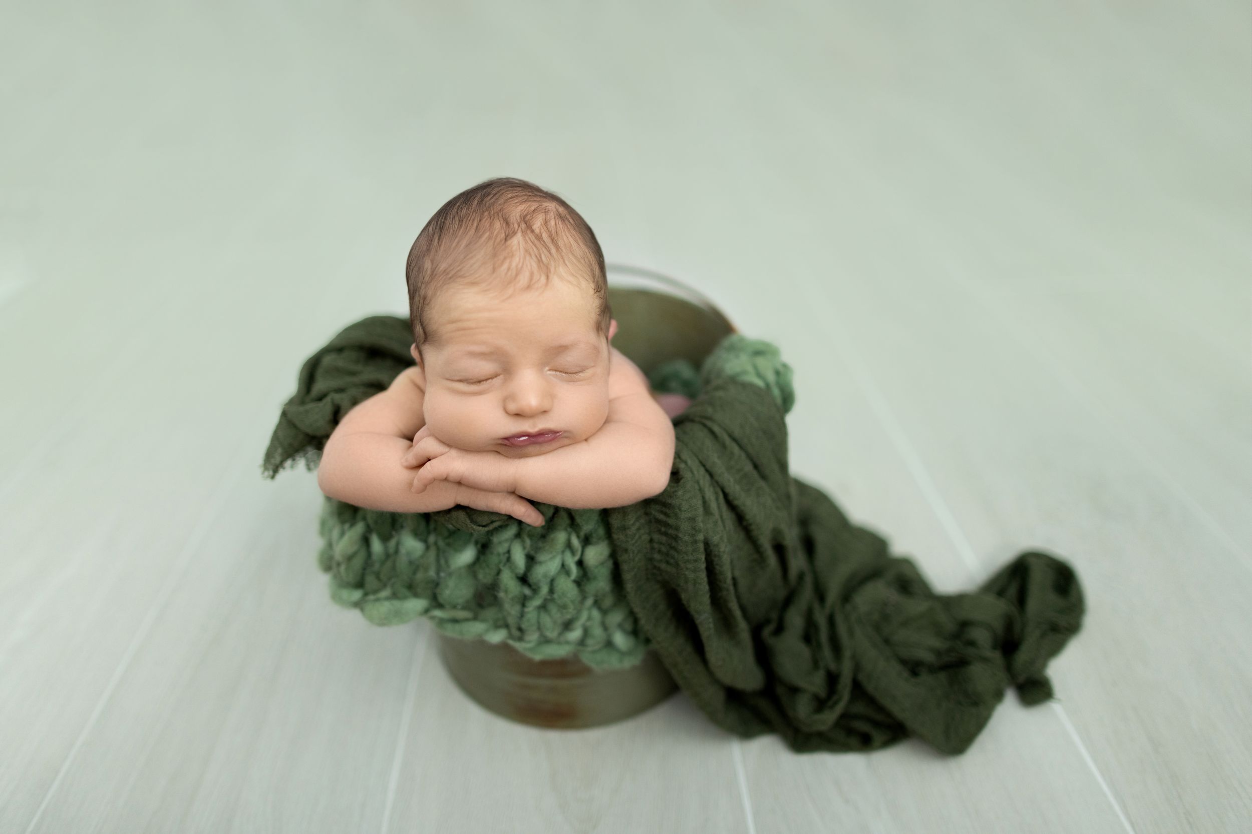 A sleeping newborn baby nestled in a green bowl, surrounded by soft fabric on a green background. Photos by Tampa Photographer DigitalMyst P